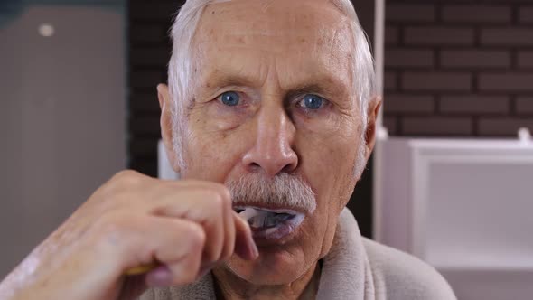 Senior Man Grandfather in Bathrobe Brushing Teeth, Looking Into Mirror. Morning Hygiene at Bathroom