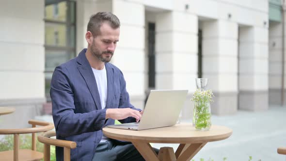 Man Pointing at Camera While Using Laptop Sitting in Outdoor Cafe