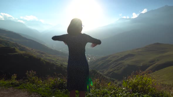 A Young Woman Stands in a Dress and Raises Her Hands Up Looking at the Mountains. The Sun Shines