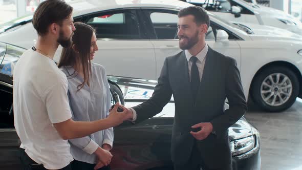 Successful Deal Between Car Salesman and Young Couple in Car Dealership.