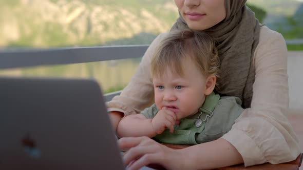 Mother Multitasking Holding Baby Infant and Using Computer Laptop at Home