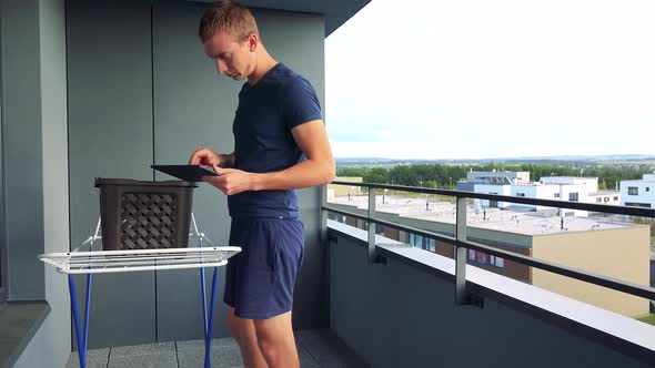A Young Man Hangs Clothes on the Drying Rack on the Balcony and Works on the Tablet
