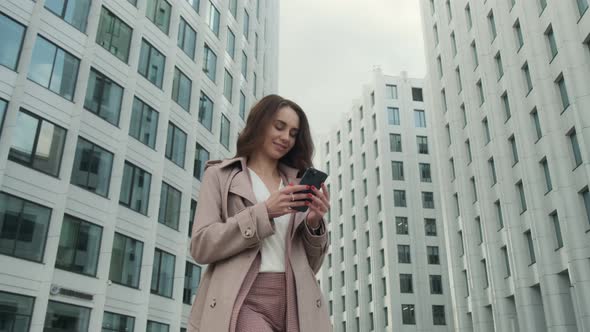 Cheerful young Caucasian woman standing in city street and texting on mobile phone with smile.