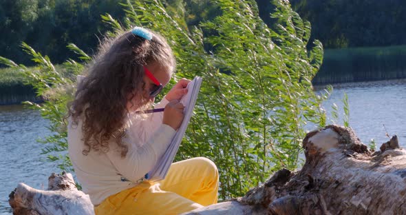 Child Is Sitting on a Log Makes the Drawing in the Open Air at Sunset.