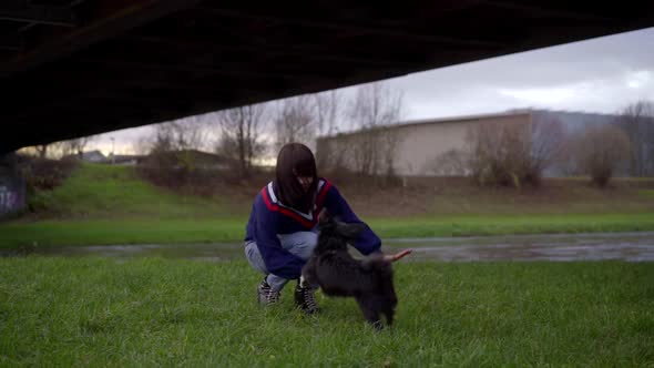 a Brunette in Jeans and a Blue Sweater Walks with Her Dog Near the River Under the Bridge. They Play