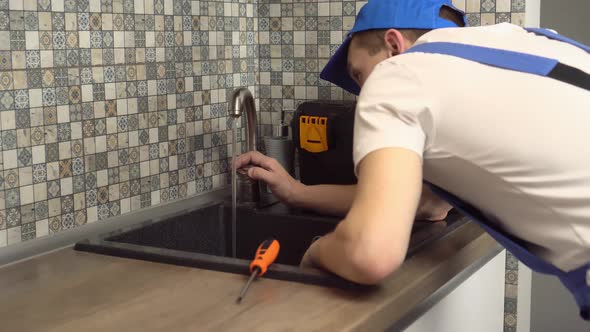 plumber in jumpsuit and white T-shirt examines faucet in kitchen in apartment.