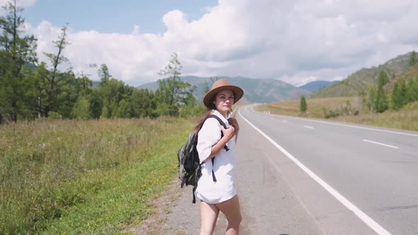 Attractive Brunette Girl in a Hat with a Backpack Travels in the Mountains Hitchhiking, Votes the