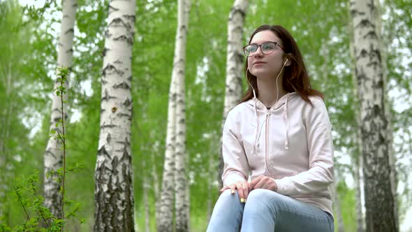 A Young Woman Sits in Nature with Headphones in Her Ears. A Girl Sits on a Stump in a Birch Forest