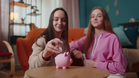 Girls Friends Siblings Sitting on Floor and Take Turns Dropping Dollar Banknote Into Piggy Bank