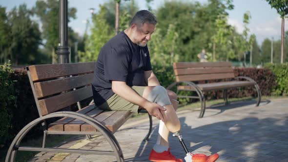 Side View Greyhaired Male Amputee Adjusting Artificial Leg Sitting on Bench in Park on Sunny Day