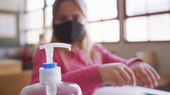 Girl wearing face mask sanitizing her hands while sitting on her desk at school