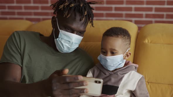 Father and Son are Sitting on a Sofa in Medical Face Masks Watching Video on Smartphone
