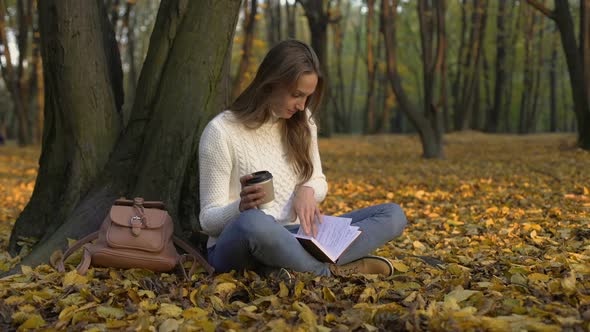 Cute Girl Sitting Under Tree in Park, Enjoying Coffee and Reading Her Diary