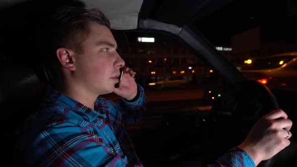 A Young Man Is Driving a Car and Talking on the Phone at Night. Driver Side View