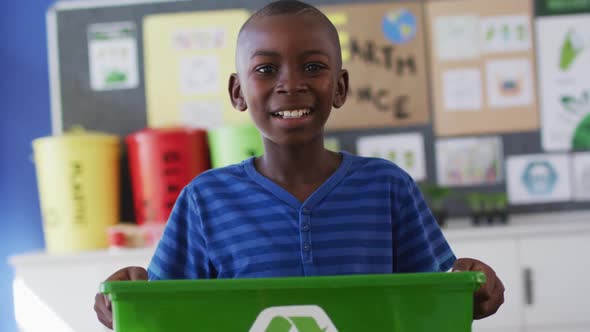 African american schoolboy smiling, holding recycling bin, standing in classroom