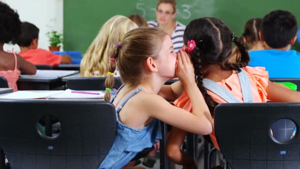 Schoolgirl whispering into her friend s ear in classroom