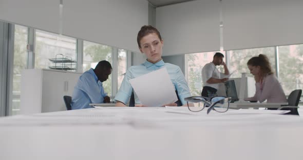 Caucasian Businesswoman Sitting at Desk Reading Document in Open Space Office