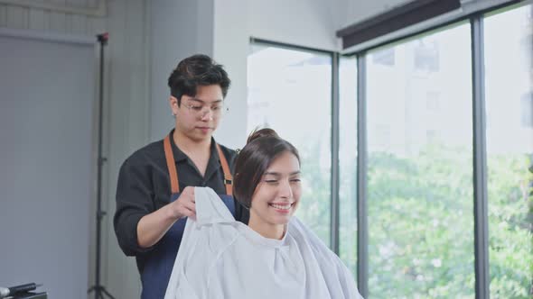 Asian professional hair stylist man puts protective cape apron to Caucasian beautiful woman in salon