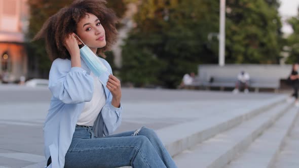 Young African American Girl Afro Woman with Curly Hairstyle Sits on Sidewalk in City Wears Medical