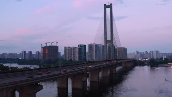 City Traffic Over River Bridge with Buildings and Cranes on Background