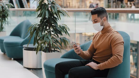 Young Man in Medical Mask Sits Indoors Typing Message on Social Network on Smartphone Sends Email