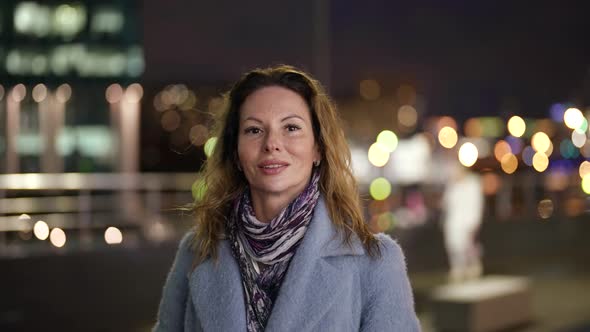 Portrait of a Smiling Blonde with Dark Eyes in a Light Coat and Scarf in the Evening Against the