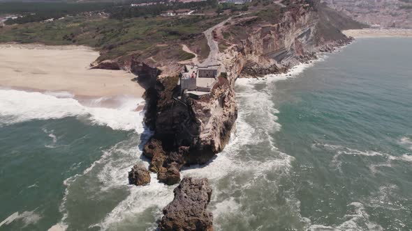 Waves crashing on rocky cliffs with Fortress of Saint Michael the Archangel and Nazare lighthouse