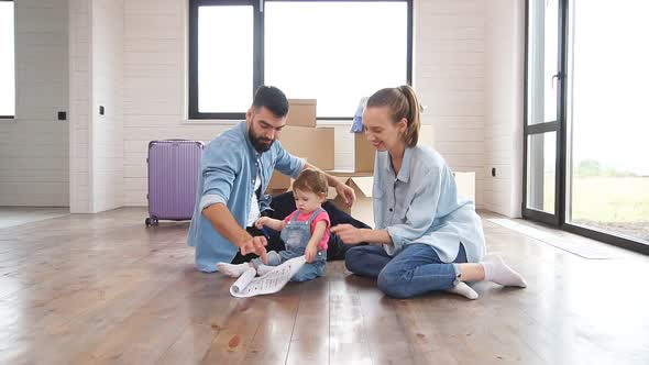 Caucasian Family of Three Sit on Floor in New House and Look at House Plan.