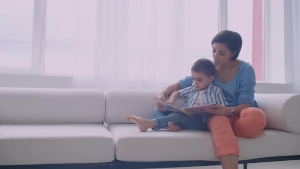 Happy Mother and Little Son Reading a Book in the Morning Together in the Living Room at Home