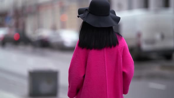 a Brunette in a Black Hat Walks Away Against the Background of a Blurry City Street Turns Around