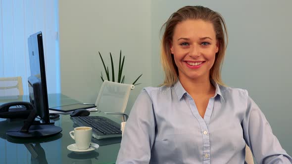 A Young, Beautiful Woman Sits at a Desk in an Office and Smiles at the Camera