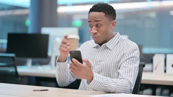 Shocked African Businessman Using Smartphone and Drinking Coffee