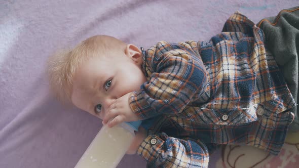 Funny Boy Eats Meal From Bottle Lying on Soft Bed with Plaid