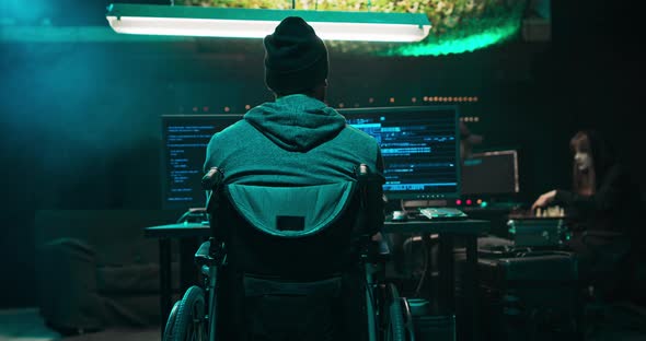 Shot From the Back to Man in a Wheelchair a Hooded Hacker Works on a