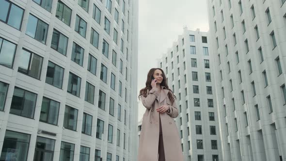 Cheerful young Caucasian woman standing in city street and texting on mobile phone with smile.