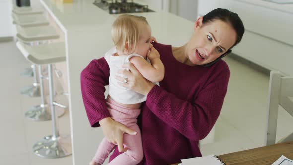 Caucasian mother holding her baby and talking on smartphone at home