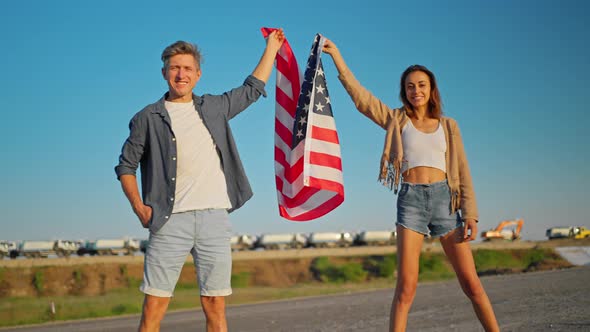 Happy Couple American Family Standing on Road at Sunset and Holding Flag of USA with Proud on Wind