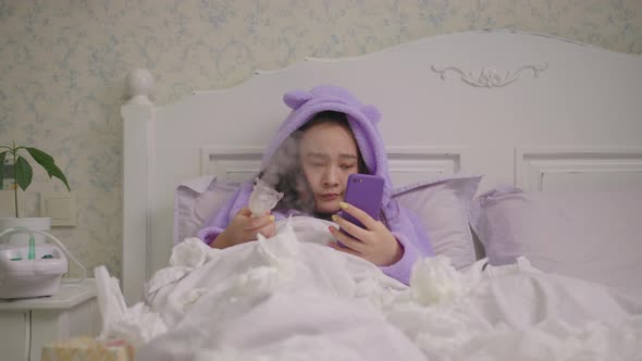 Millennial Sick Asian Woman Using Mobile Phone Instead of Using Inhaler for Flu Therapy Lying in Bed