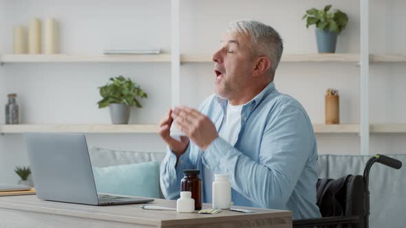 Disabled Senior Man In Wheelchair Sneezing In Handkerchief At Home