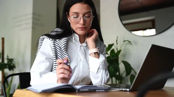 Serious young brunette woman in eyeglasses working on laptop and writing in notepad