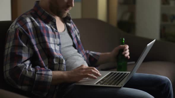 Untidy man on sofa using laptop and drinking beer, browsing internet, loneliness