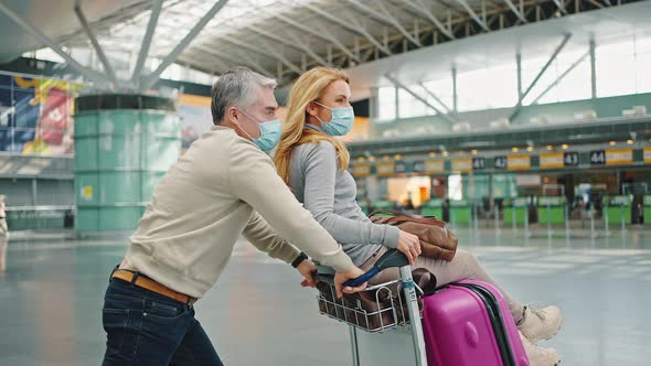 Happy Middle Aged Spouses Fooling Around at Airport Building Making Ride Race on Luggage Cart