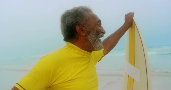 Front view of happy active senior African American man with surfboard standing on the beach 4k