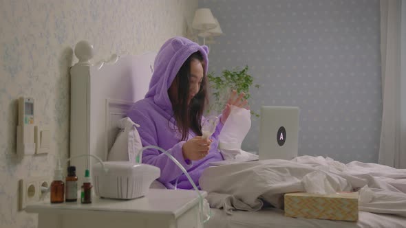 20s Sick Asian Woman Talking to Doctor Using Laptop Holding Inhaler in Hands Lying in Bed