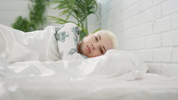 Young Beautiful Blonde Woman with Short Haircut Wakes Up in Morning at Home on Silk White Sheets