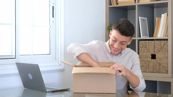 Smiling man wearing glasses unpacking awaited parcel, looking inside, sitting at work desk