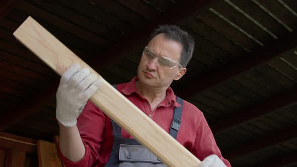 Carpenter in Protective Glasses Holds a Wooden Beam in His Hands and Examines It