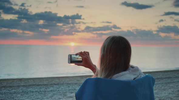 Woman Making Video of Sunset with Smartphone Sitting on the Seashore