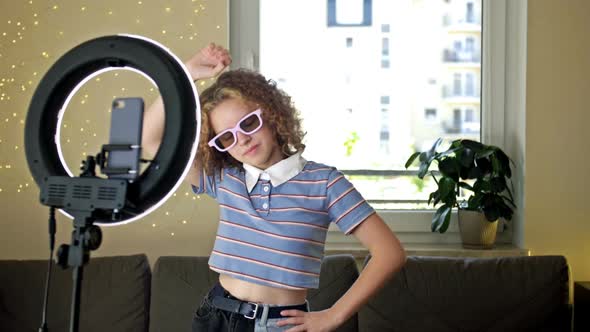 Cheerful Teen Girl Dancing at Camera Filming Video Using Phone on Tripod at Home Creating Her Trendy