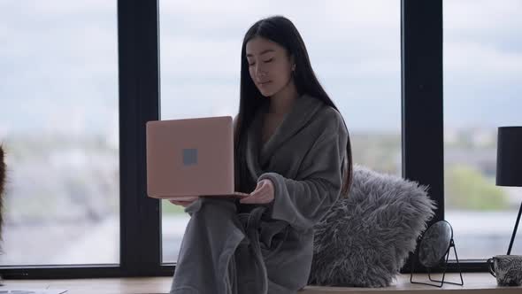 Young Slim Asian Woman in Bathrobe Sitting Down on Windowsill in Slow Motion and Typing on Laptop